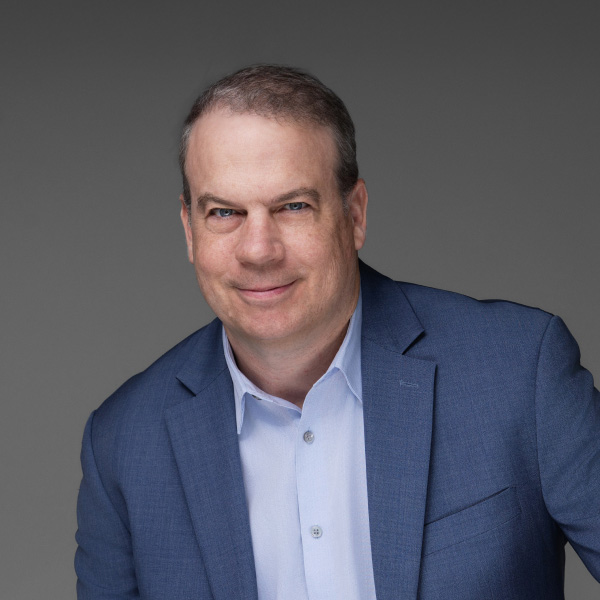 A man in a blue suit and white shirt poses with a calm and confident expression against a gray background. His posture is relaxed yet professional, conveying an approachable and competent demeanor. The lighting creates a soft contrast, highlighting the man's friendly smile and assured presence.