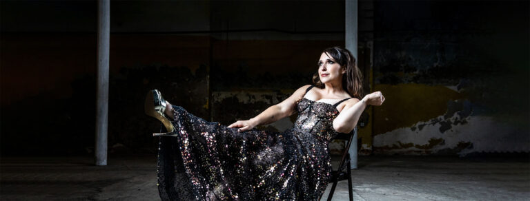 Jessica Vosk seated in a chair looking up and pointing one of her legs up