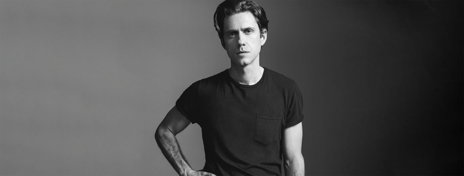Aaron Tveit, standing up, looking serious in black and white color