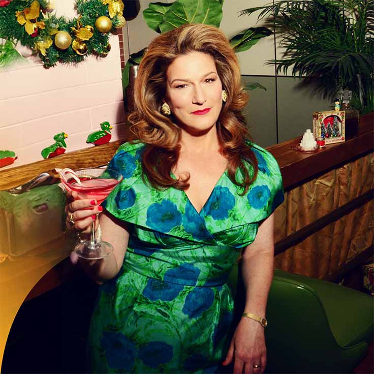 A woman with a green dress and christmas decorations in the background. She has a sugar and booze drink on her right hand