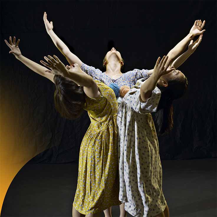 Three female dancers with their hands towards the sky, with a black background.