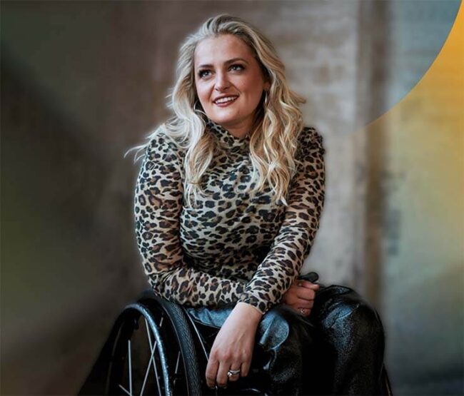 White female on a wheelchair, wearing a leopard printed shirt and with wavy blond hair, smiling
