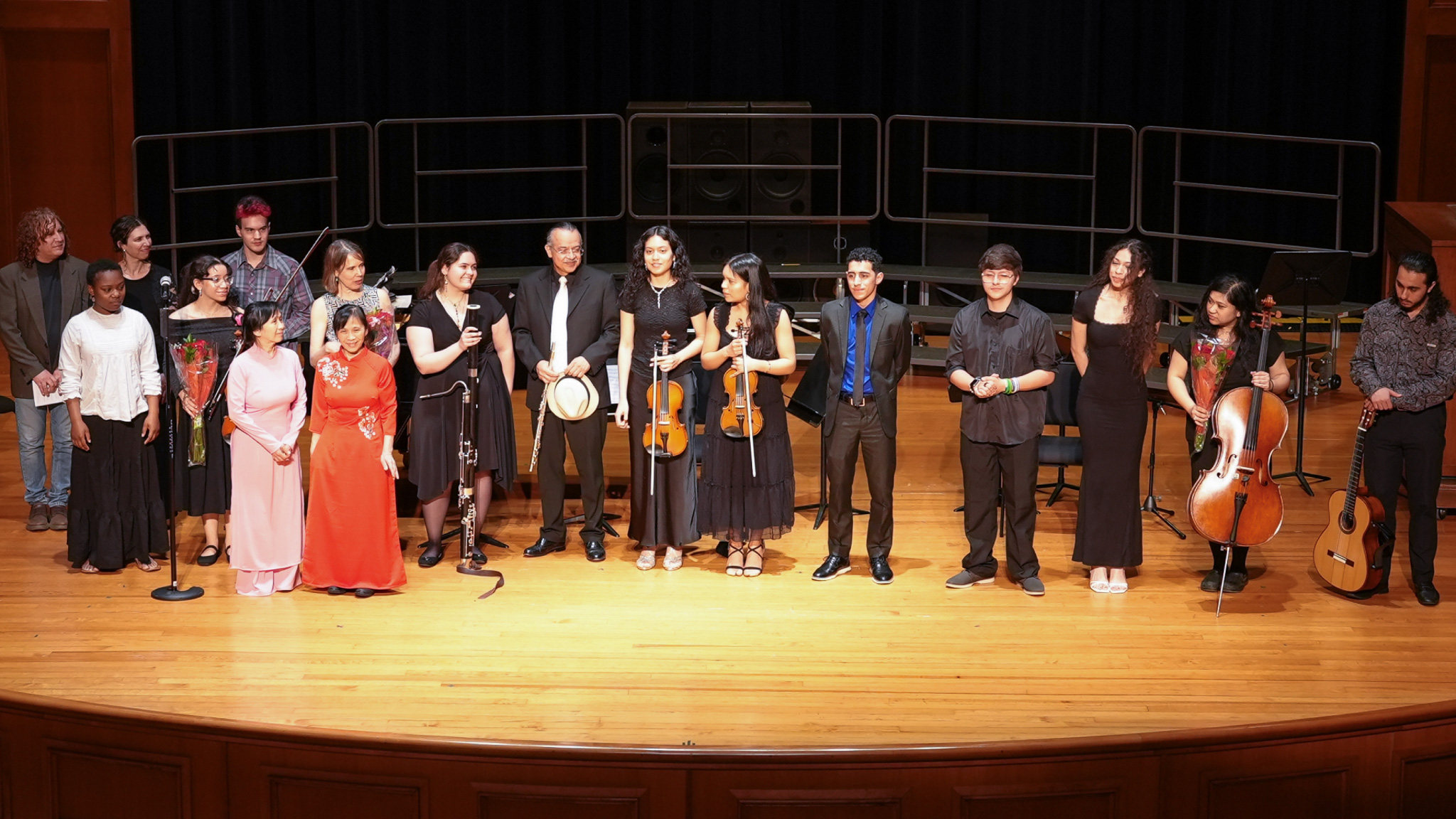 A group of musicians, dressed in formal attire, stand in a line on a concert hall stage, some holding musical instruments like a cello, violin, and flute. The diverse ensemble appears to be acknowledging an audience, set against a backdrop of choir risers and a polished wooden floor