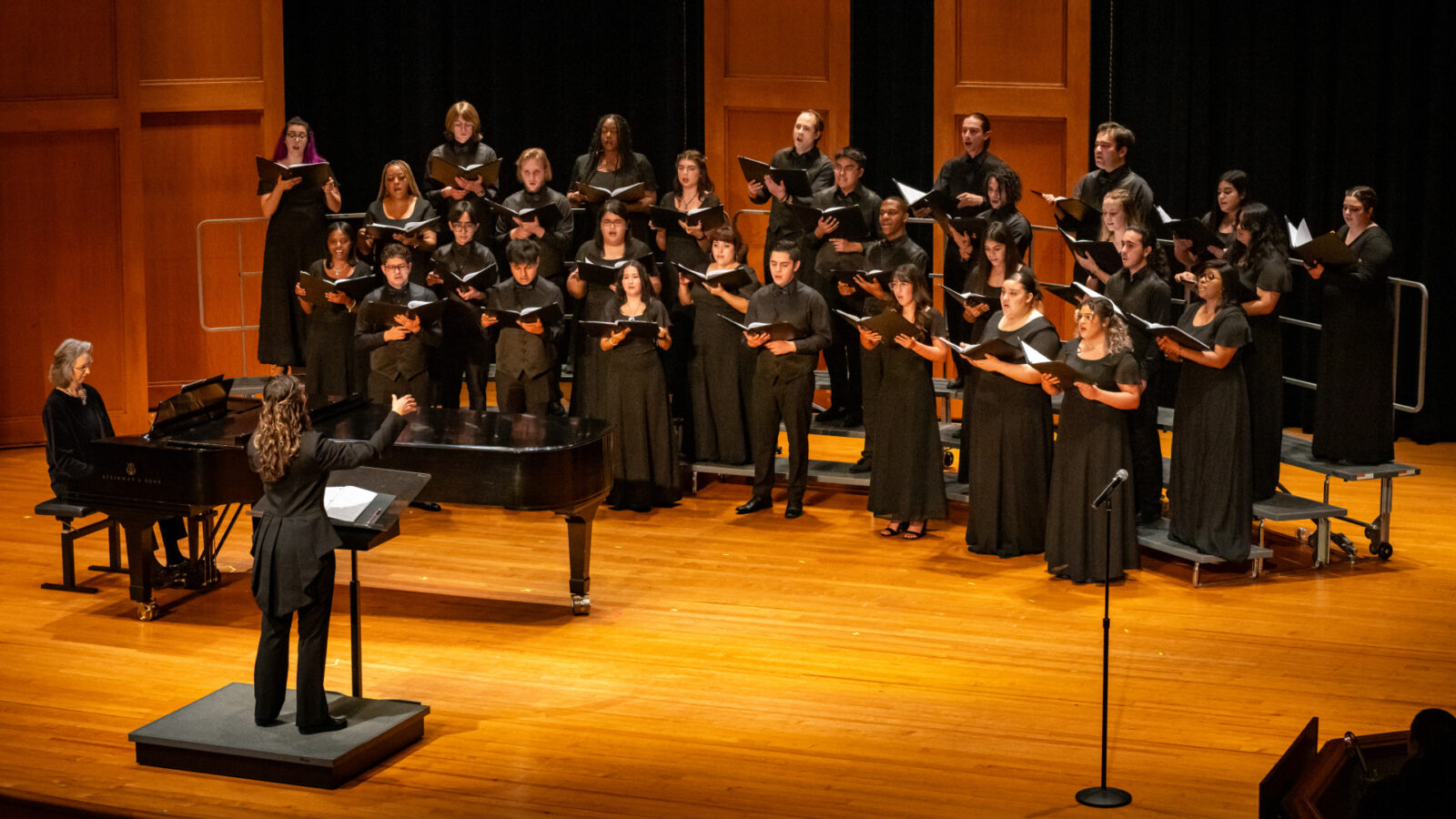 A choir performs on a wooden stage with a conductor leading the group. The choir members are dressed in formal black attire, standing in arranged rows, holding sheet music. A pianist accompanies the choir from a grand piano on stage. The performance takes place in a warm, wood-paneled concert hall, providing an elegant setting for the musical event.