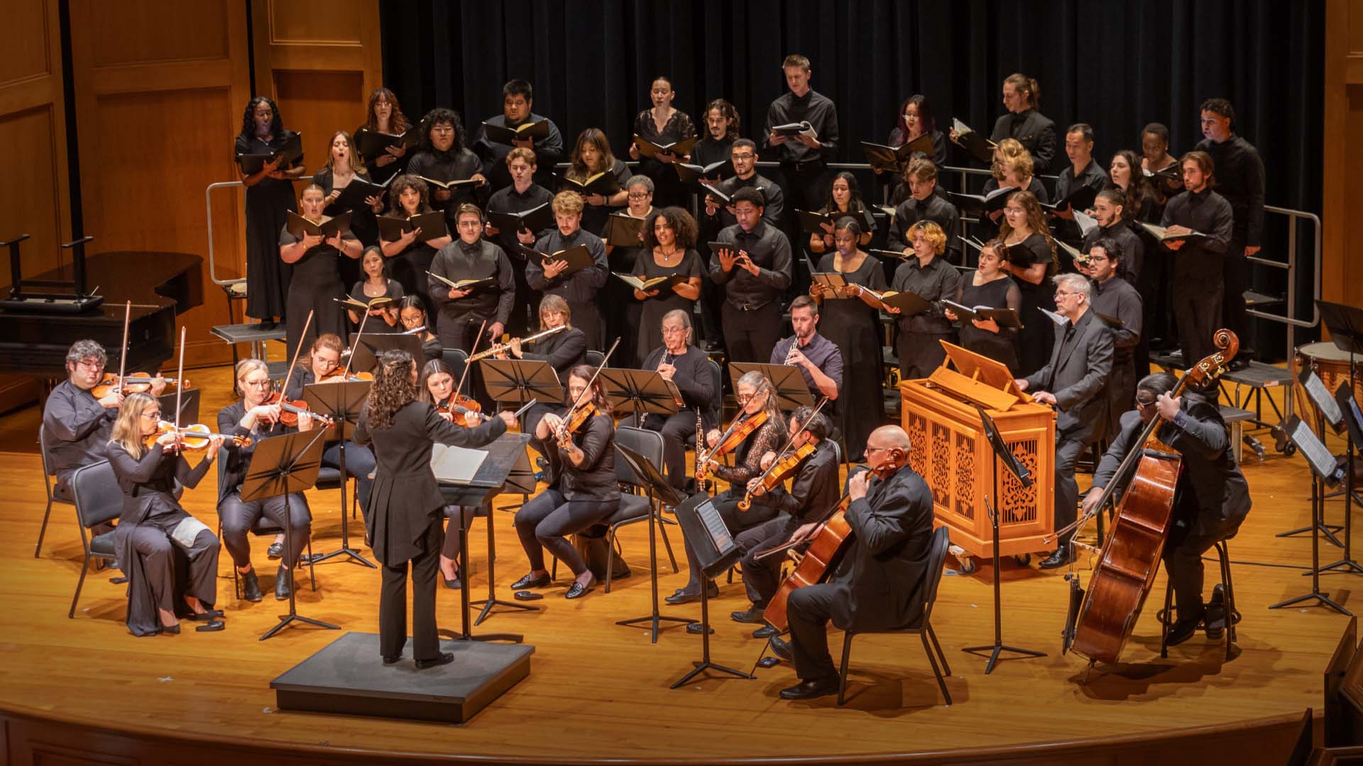 An orchestra and choir perform together on a stage. The orchestra includes string players with violins and cellos, as well as a conductor leading the ensemble. The choir, composed of numerous singers, stands elevated behind the orchestra, holding sheet music. The performance setting features warm lighting and a wooden stage