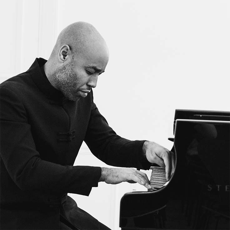 Aaron, a male with thick eyebrows, a severe expression, dressed in an all-black suit, stands next to a piano and against a white background.
