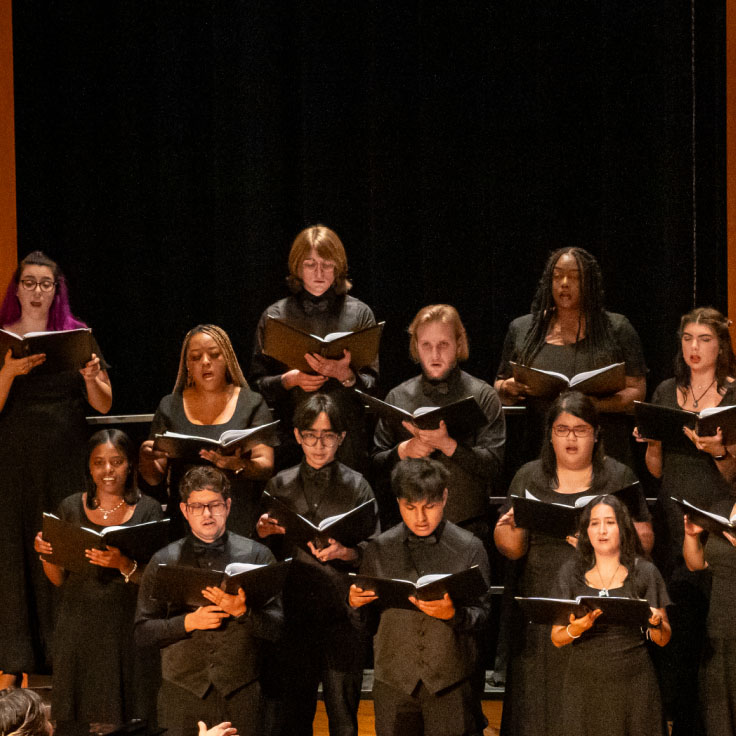 A choir performs on stage with a conductor standing in front of them and a pianist accompanying on a grand piano. The singers, dressed in formal black attire, hold sheet music as they sing in a semi-circle arrangement on risers. The stage is warmly lit with a wooden floor.