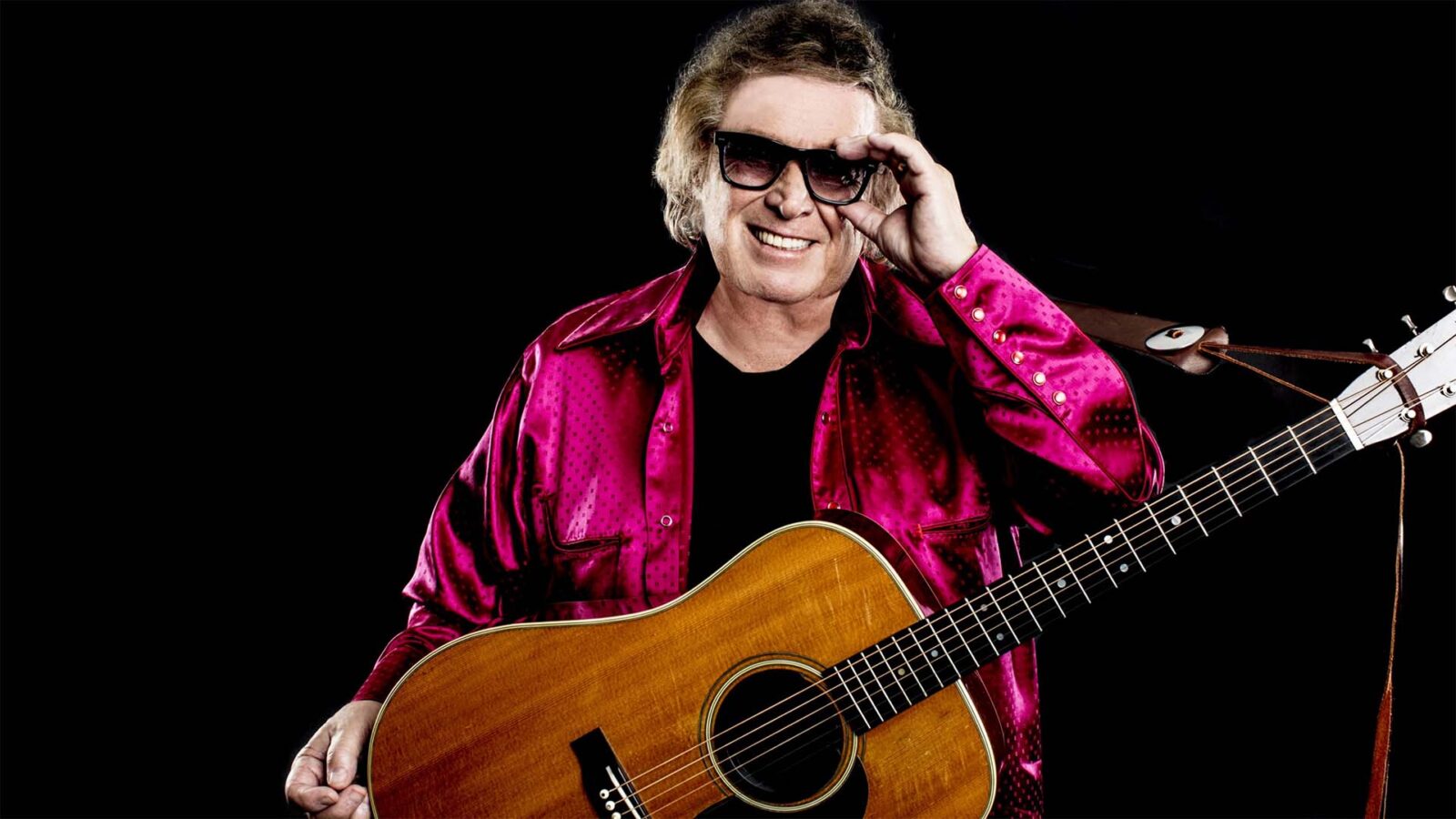 Don McLean wearing glasses, smiling at the camera with a guitar hanging from his shoulder and wearing a bright pink shirt