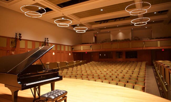 Inside the Enlow Recital Hall. Shot from the stage, showing a piano on the left and the theatre seats and lamps on the background