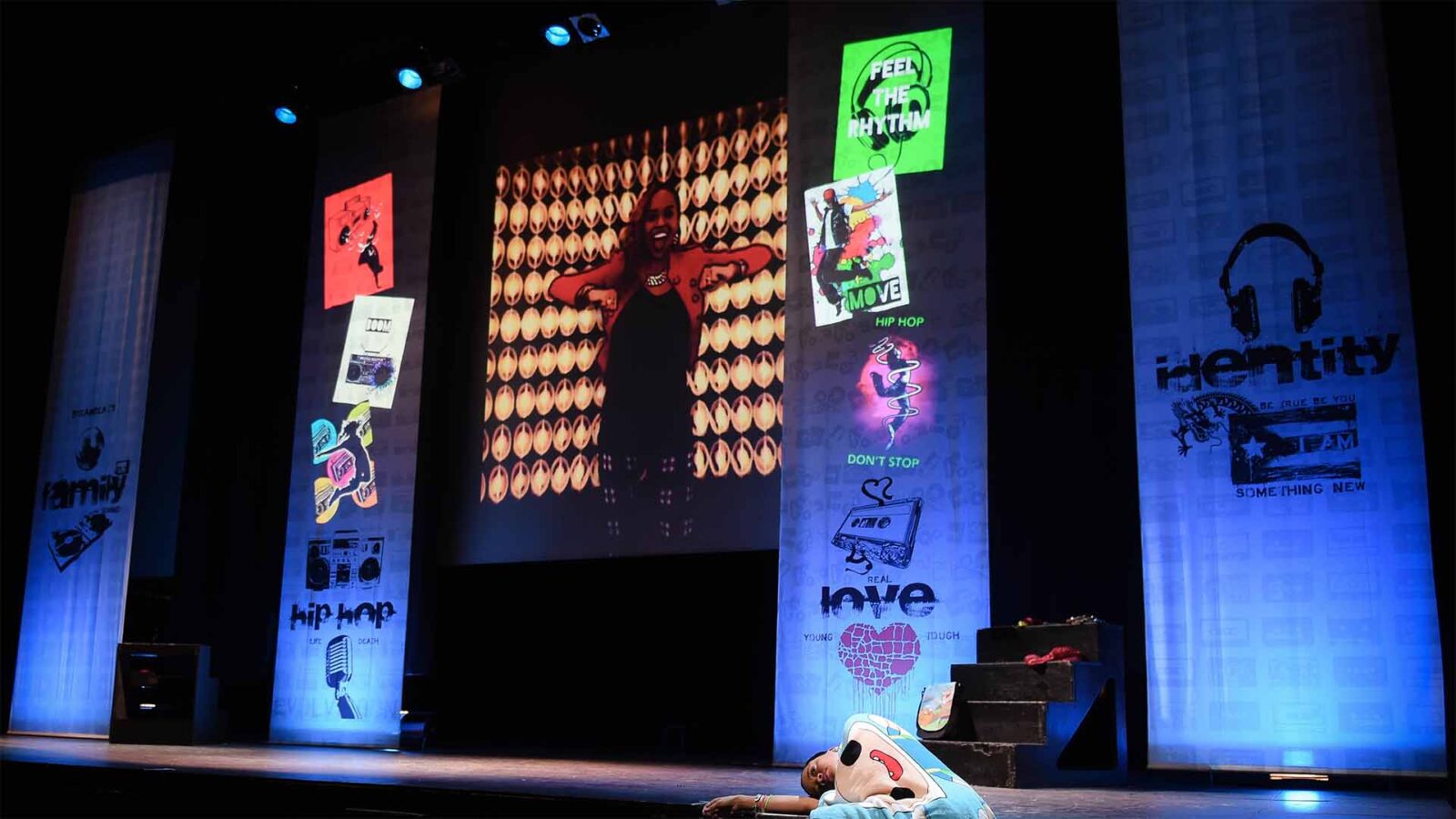 A whole stage filled with images of Cuba and dancers projected on a screen. On the stage, a woman lays on the floor with a blanket.