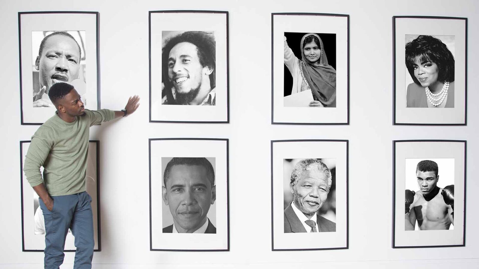 A man in a grey shirt and jeans leans on a wall of photographs of various celebrities as if showcasing them.