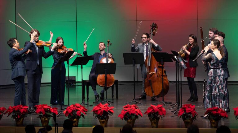 A group of musicians on stage with their violins and other wind instruments. A gradient of green and red fills the background for a holiday feel.
