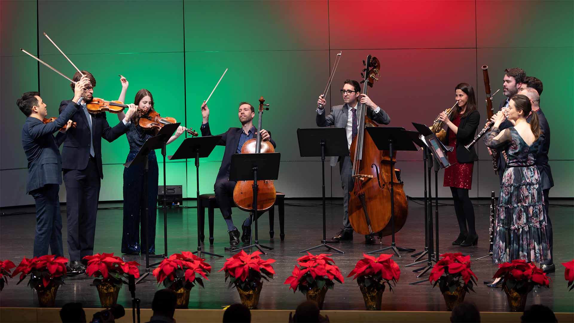 A group of musicians on stage with their violins and other wind instruments. A gradient of green and red fills the background for a holiday feel.