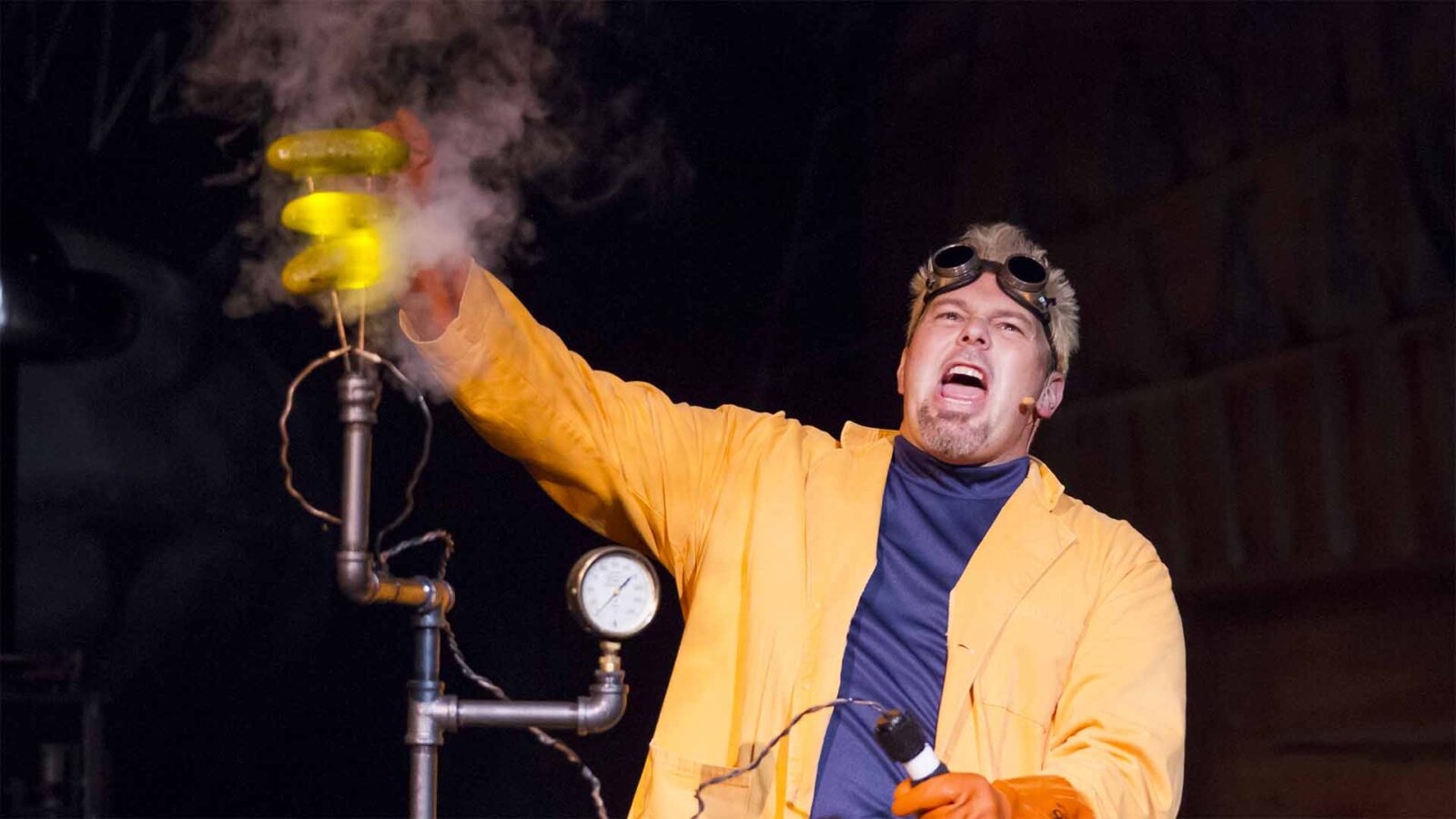 Doktor Kaboom dressed in a yellow coat, with goggles on his head and pointing to a glass tube filled with smoke as if his scientific experiments took a wrong turn.