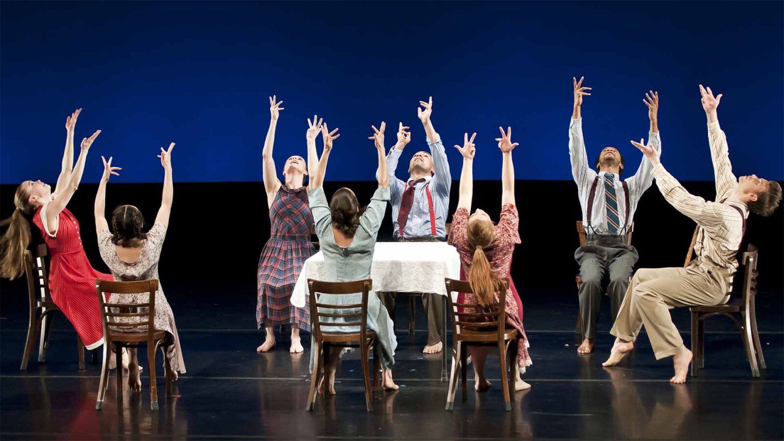 A group of dancers on stage dressed differently. They are sitting down with their arms towards the ceiling.