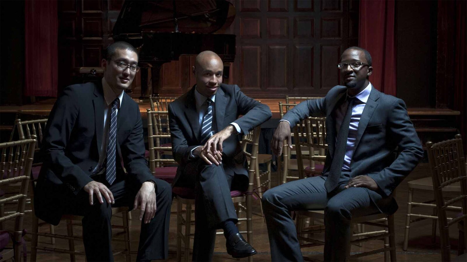Three gentlemen in suits are sitting and looking at the camera. At their back, a black piano on a stage appears.
