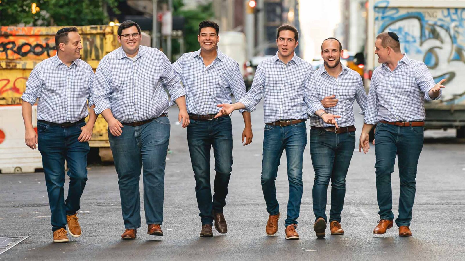 Six men dressed in jeans and gray shirts are standing in the streets of NYC and looking like they are walking towards the camera, laughing and talking to each other.