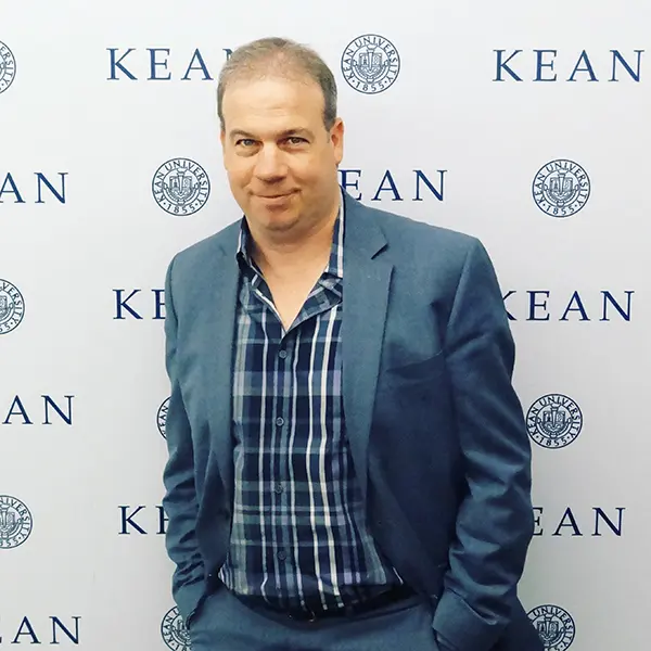 A white man, dressed in a blue suit, with a Kean logo background