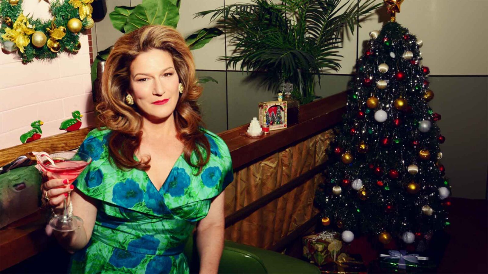A redhead woman in a green silk dress sits by a Christmas tree with a drink in her hand.