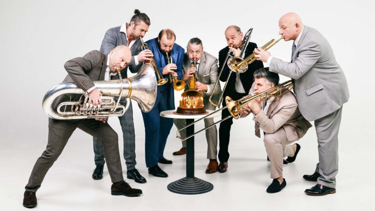 7 brass players of Mnozil Brass using their trumpets to blow a chocolate cake with the number 30 on it.