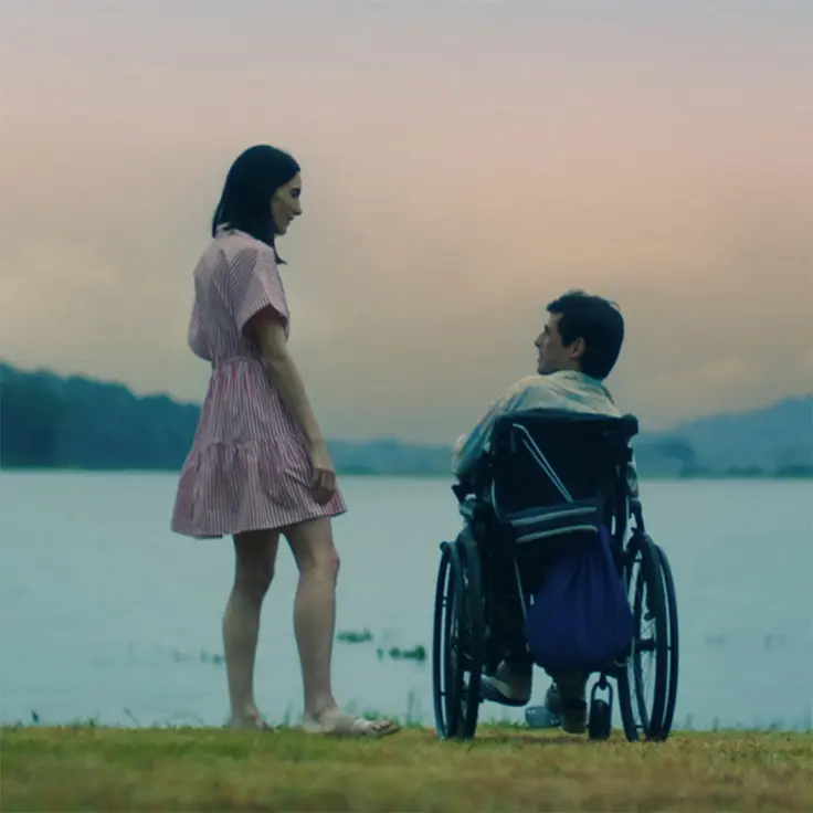 Two people by a lakeside at sunset, with one person standing on the shore and another seated in a wheelchair facing the water.