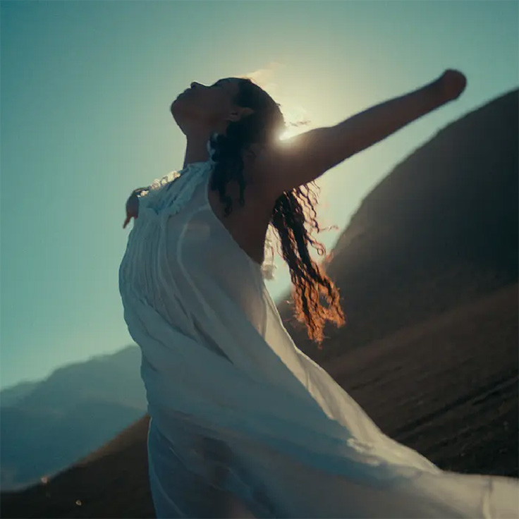 A woman in a white flowing dress stands with arms outstretched, silhouetted against the sun. Her pose and the mountainous backdrop evoke a sense of freedom, tranquility, and connection to nature. The lighting emphasizes her silhouette and the movement of her dress, symbolizing liberation and serenity.