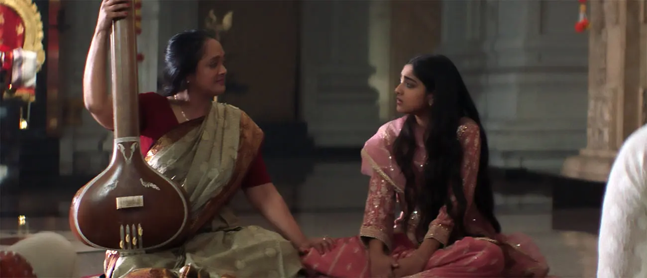 Two people seated on the floor of an ornate interior, with one holding a large stringed instrument and the other wearing a pink traditional outfit, facing each other in conversation.