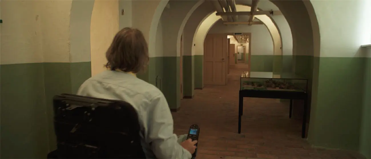 Person using a powered wheelchair moves through a long arched hallway with tiled floors and display cases.