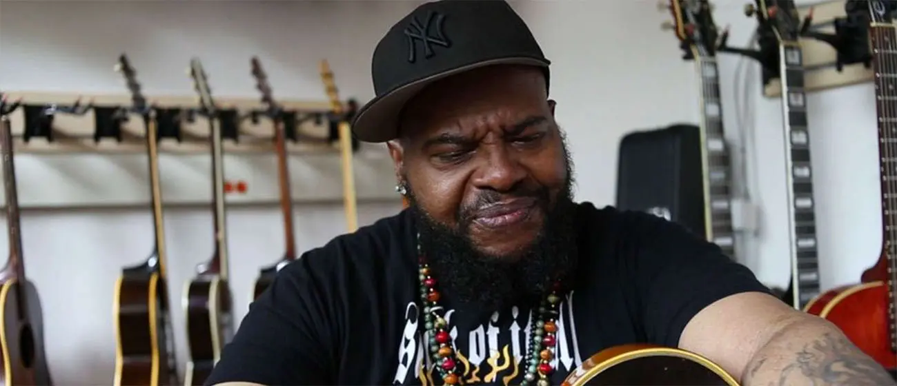 Musician wearing a black cap and beaded necklace sits among several guitars, holding an acoustic guitar in a recording or rehearsal space.