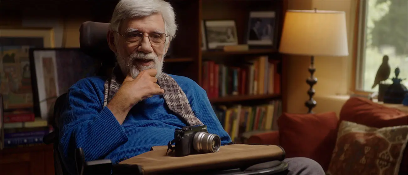 Person seated in a wheelchair with a camera on their lap in a cozy room filled with bookshelves, framed artwork, and warm lighting.