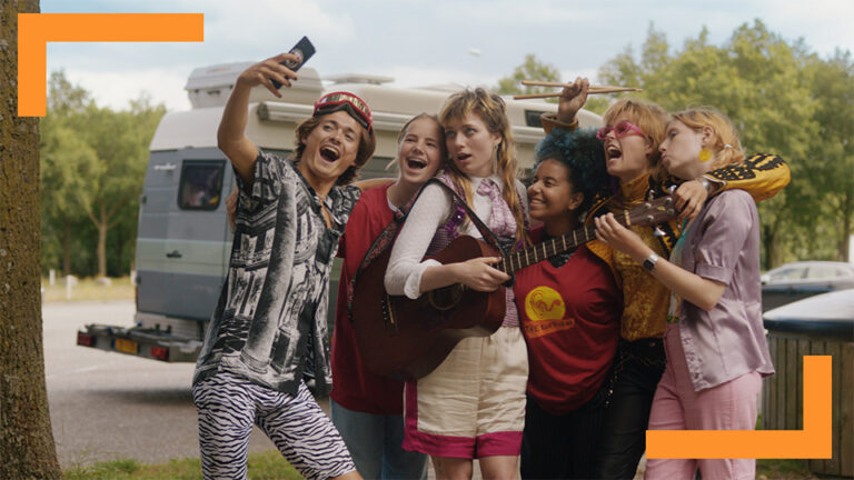 A group of six friends, diverse in appearance, joyfully gather for a selfie. They are outdoors next to a camper van, suggesting a road trip adventure. One person is holding a guitar, adding a musical element to the scene. The lively expressions and casual attire capture a moment of carefree fun and friendship, set against a rural backdrop with trees.