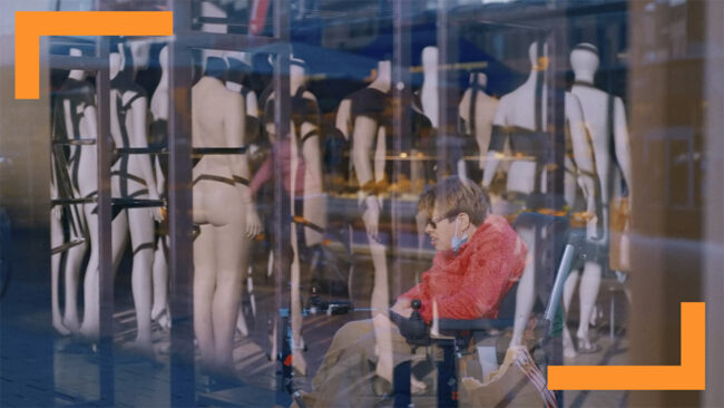 A window reflection composition featuring a person in a red jacket using a wheelchair juxtaposed with a display of mannequins. The image blends different layers of reality and reflection, creating a dynamic urban scene that juxtaposes human presence with commercial display. The visual contrast highlights themes of accessibility, societal norms, and the intersection of consumer culture with everyday life.