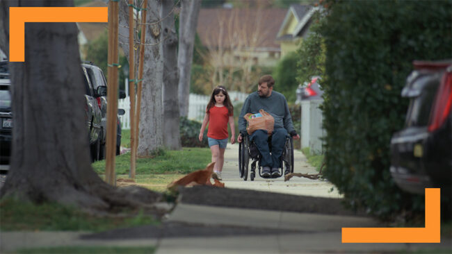 A man in a wheelchair accompanies a young girl walking alongside him on a suburban sidewalk, while a dog walks on the grass nearby. The scene exudes a sense of everyday life, accessibility, and family bonding, highlighted by the springtime setting and the presence of greenery along the path. The image captures themes of mobility, companionship, and the warmth of shared moments in a community environment.