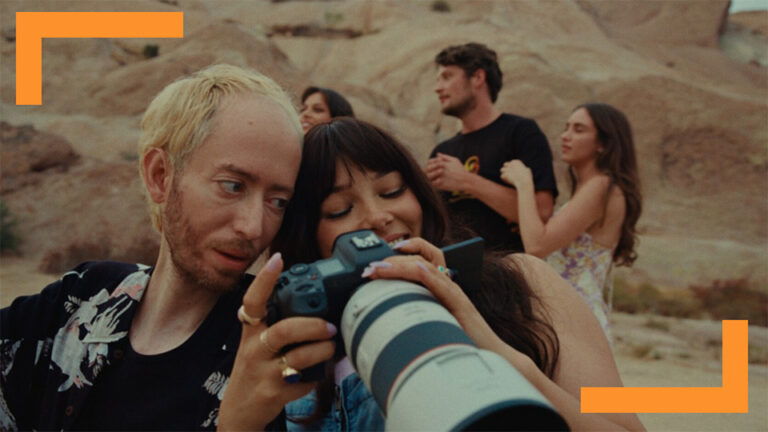 A group of friends gather in a desert landscape, with two individuals in the foreground closely examining a photograph on a camera. They display curiosity and excitement as they bond over photography. The setting is outdoors, showcasing natural rock formations and a warm ambiance, emphasizing friendship, creativity, and exploration in nature.