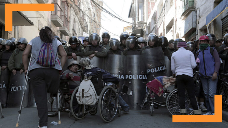 A group of protesters with disabilities, some in wheelchairs and others with crutches, confront a line of riot police. The scene unfolds in an urban street, reflecting themes of resistance and civil rights activism. The police, equipped with helmets and shields, stand in a defensive line, creating a barrier. The image captures the struggle and determination of marginalized individuals fighting for their rights against authoritative forces.