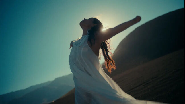 A woman in a white flowing dress stands with arms outstretched, silhouetted against the sun. Her pose and the mountainous backdrop evoke a sense of freedom, tranquility, and connection to nature. The lighting emphasizes her silhouette and the movement of her dress, symbolizing liberation and serenity.
