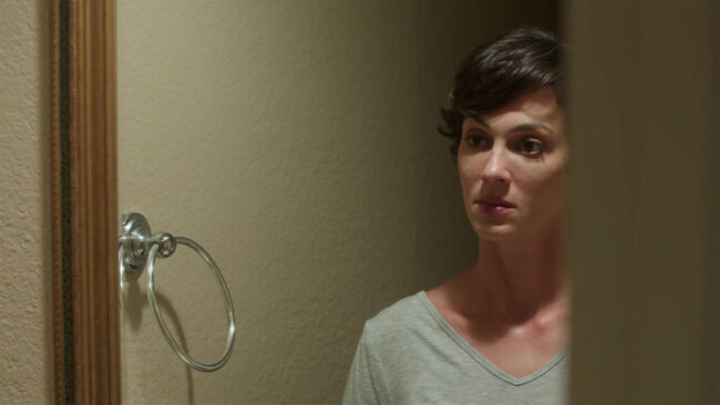 A woman with short dark hair stands in a dimly lit room next to a mirror, reflecting a contemplative and introspective mood. The wall's neutral tone contributes to the subdued and thoughtful atmosphere.