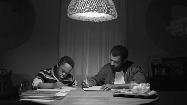A black and white image showing a father and son engaged in study at a dining table, illuminated by a single overhead light.