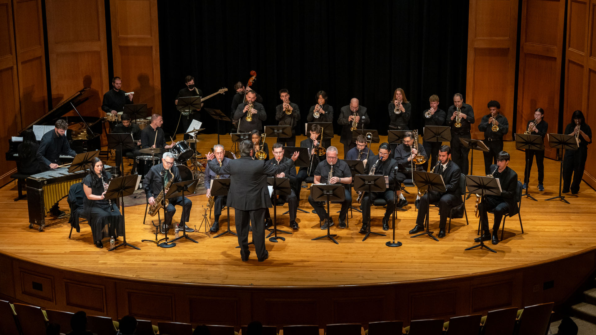 A large orchestra performs on a wooden stage in an auditorium. The conductor stands at the forefront leading various musicians playing a range of wind and percussion instruments. The musicians, dressed in formal attire, are deeply engaged in the performance under warm stage lighting. The background features an elegant, dark curtain, and the ambiance suggests an evening of classical music.