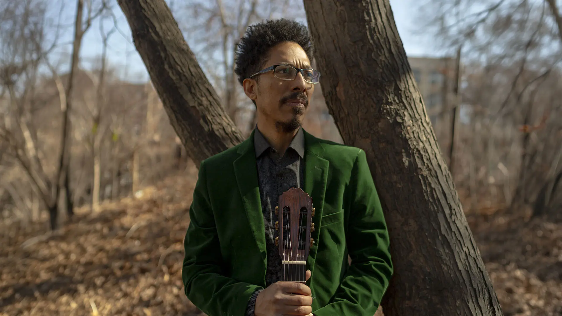 A musician wearing a green velvet jacket and glasses stands contemplatively against a tree, holding a guitar. The setting is a serene outdoor environment with bare trees and soft sunlight, suggesting late autumn or early winter.