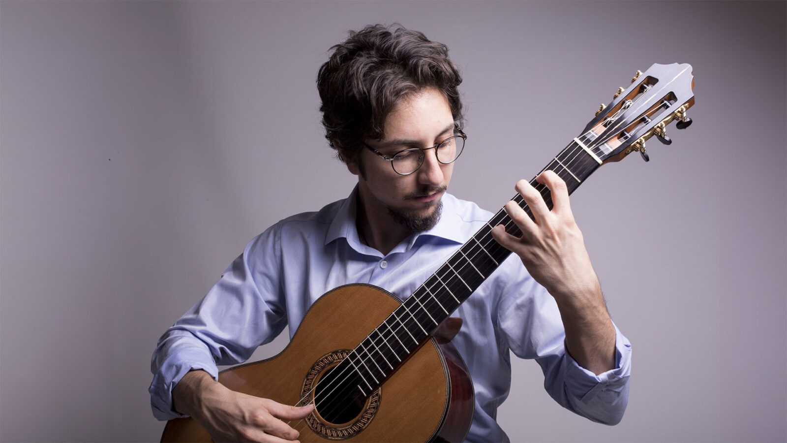 A man wearing glasses and sitting down and playing a guitar