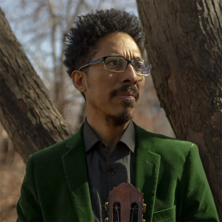 A musician wearing a green velvet jacket and glasses stands contemplatively against a tree, holding a guitar. The setting is a serene outdoor environment with bare trees and soft sunlight, suggesting late autumn or early winter.
