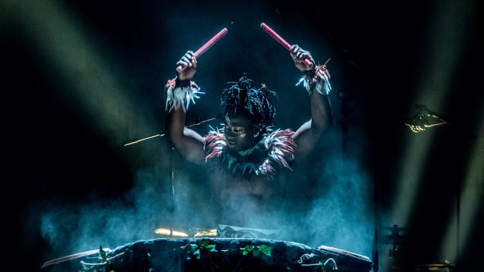 A drummer, illuminated dramatically on stage, is captured mid-performance. The drummer is shirtless, wearing decorative cuffs made of feathers around his wrists, and has intricate face paint. He holds two red drumsticks high above his head, poised to strike the large drum in front of him. The background is dark and misty, with beams of light cutting through the haze, adding to the intense and dynamic atmosphere of the scene.