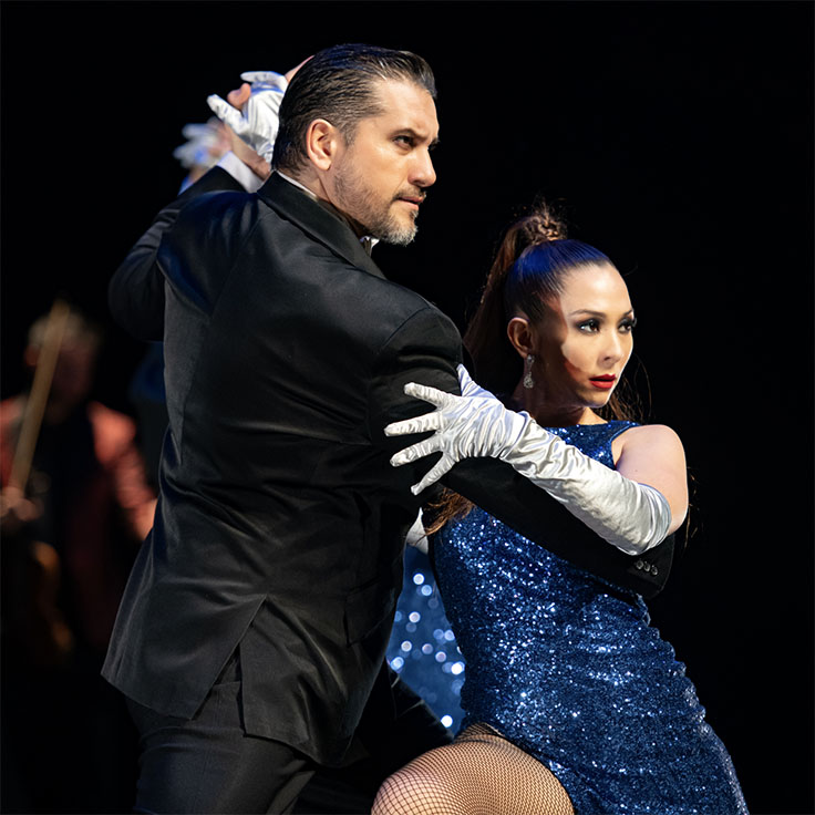 A man and a woman are captured mid-dance in a dramatic tango pose. The man, dressed in a sleek black suit, holds the woman firmly, showcasing his strong and focused expression. The woman, wearing a sparkling blue dress with long, white gloves, has her hair styled in a high ponytail and looks intensely at her partner.