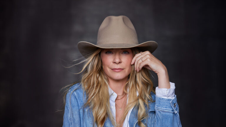 LeAnn with long, wavy blonde hair, wearing a wide-brimmed beige hat. She is dressed in a denim jacket over a white blouse. Her right hand is gently touching the brim of her hat, while her left hand rests near her face, showcasing a delicate ring on her finger. She gazes directly at the camera with a calm and confident expression. The background is dark and blurred, highlighting her as the focal point of the image.