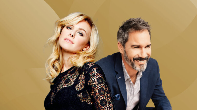 A man and a woman are posing against a light golden background. The woman, on the left, has long, wavy blonde hair and is wearing a black lace top, leaning slightly towards the man. She has a confident and relaxed expression. The man, on the right, has short, graying hair and a beard, and is wearing a navy blazer over a light-colored shirt. He is smiling warmly and looking slightly off to the side. Both appear to be relaxed and enjoying the moment.