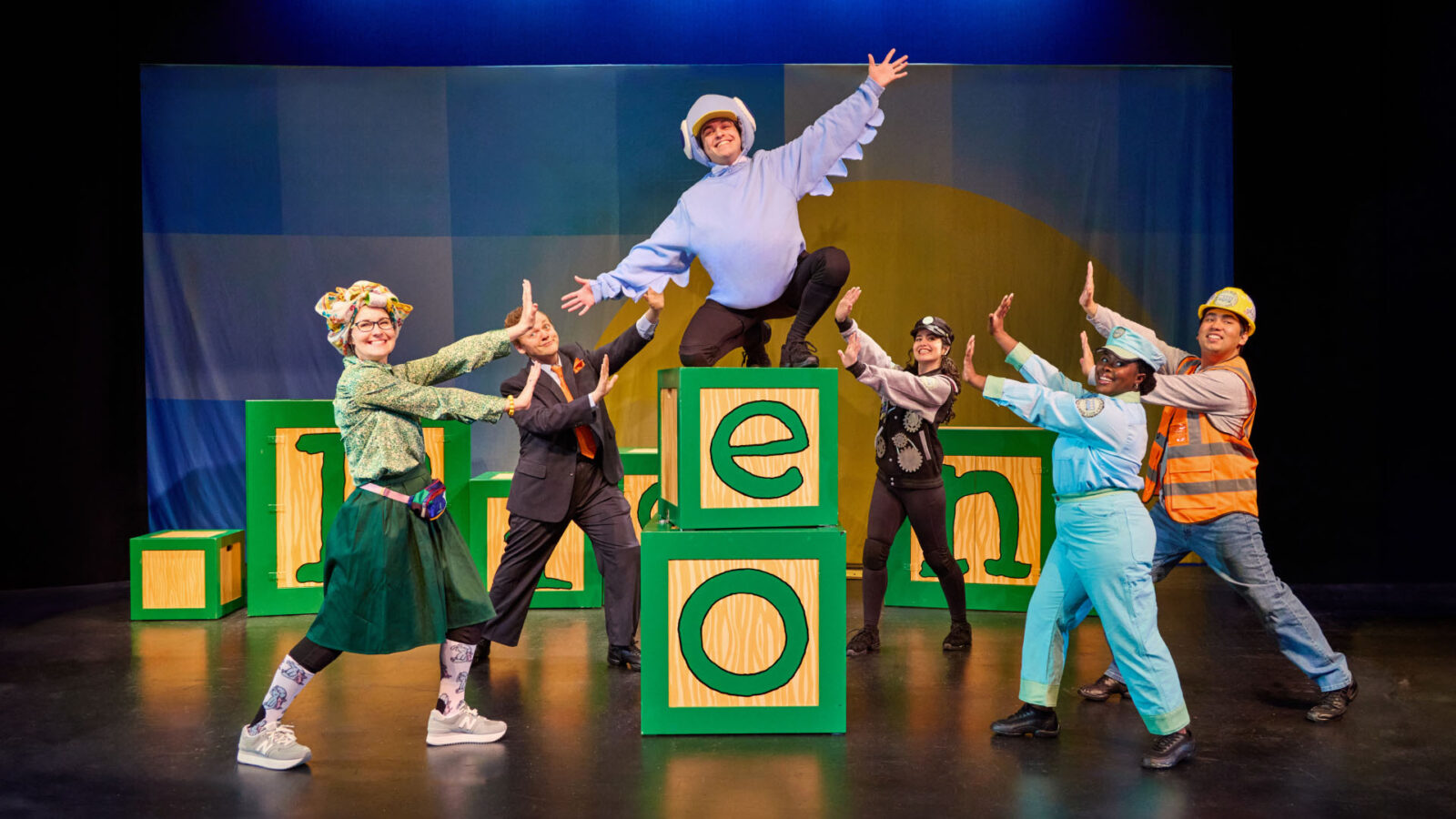 A group of six performers is on stage, striking a dynamic and playful pose. The central figure is dressed in a light blue costume with a helmet, resembling a pigeon, and is perched on large green blocks with the letters "e" and "o." Surrounding the central figure are five other performers, each dressed in colorful and whimsical outfits, including a character in a construction worker's vest, a person in a light blue uniform, and others in various playful costumes. The backdrop features a simple, colorful design, enhancing the cheerful and energetic atmosphere of the performance.