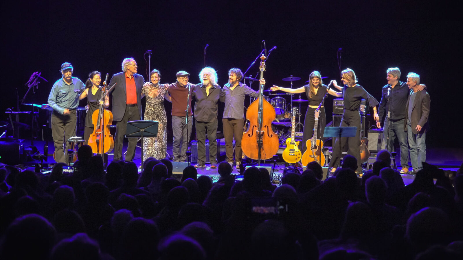 A group of eleven musicians stands on stage, arm in arm, taking a bow before an audience. The performers, a mix of men and women, are dressed in a variety of outfits, from casual to formal. Several instruments, including guitars, a cello, and a double bass, are visible on stage along with microphones and other musical equipment. The background is dark, with stage lights illuminating the performers, creating a warm and appreciative atmosphere as the audience watches.