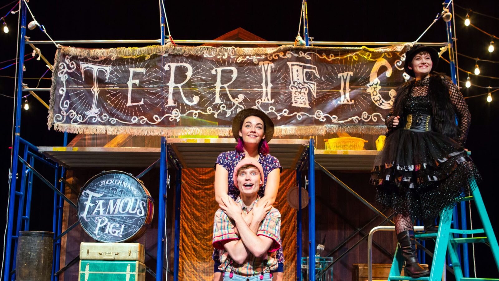 Three performers are on stage in a whimsical setting inspired by "Charlotte's Web." The central figure, a young man with a pink beanie, is kneeling and looking up with an excited expression. Behind him stands a young woman with pink hair, wearing a straw hat and a floral dress, smiling warmly. To the right, a woman dressed in a black, gothic-style outfit with a netted top, a fur vest, and a layered skirt, stands confidently on a ladder. Above them is a large banner with the word "TERRIFIC" written in a playful, artistic font, and nearby is a drum labeled "Zuckerman's Famous Pig!" The stage is decorated with colorful lights and props, creating a vibrant and lively atmosphere.