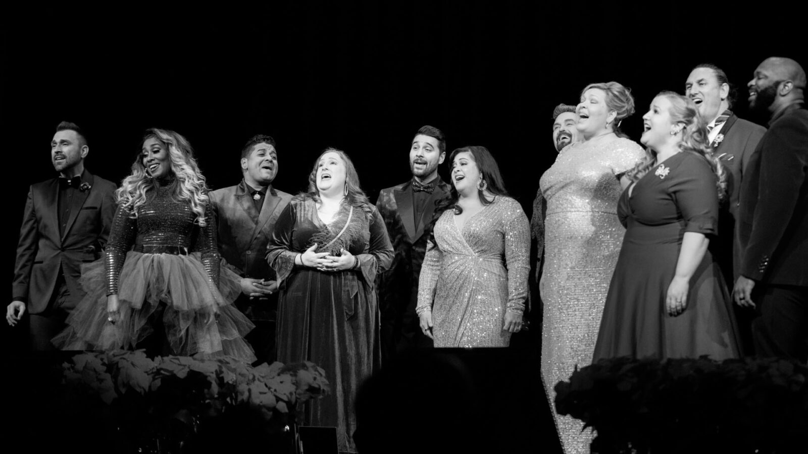 A group of eleven singers stands on stage, captured in a black and white photo. They are arranged in a line, with expressions of joy and passion as they perform. The women are dressed in elegant dresses, some with sequins and some with tulle, while the men wear sharp suits. The stage background is dark, emphasizing the performers who are brightly lit, creating a striking contrast. The audience is partially visible in the foreground, watching the performance attentively.